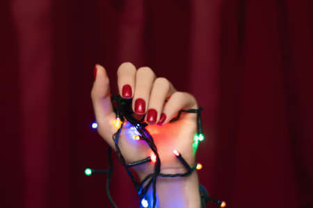 Female hand with red nail design. Glitter red nail polish manicure. Woman hand hold Christmas garland on dark red fabric background. Holiday manicure concept.の写真素材