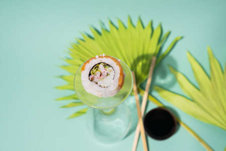 Sushi roll on bottom of wine glass with chopsticks and soy source decorated with green tropical leaves on green background. traditional Japanese food. Restaurant food, cafe concept.の写真素材