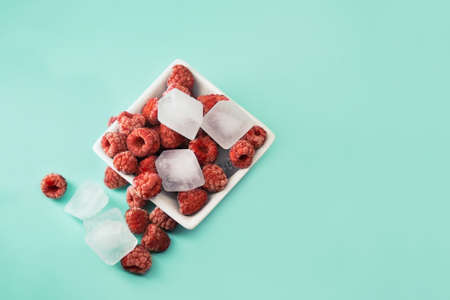 Frozen raspberries in white plate on green background. Frozen ripe raspberries with ice cubes. Whole raw frozen berries.の写真素材