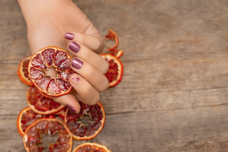 Female hand with crimson nail design. Glitter crimson nail polish manicure with star nail art. Woman hand hold dry oranges on wooden background. copy space.の写真素材