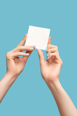 Female hands with blue manicure holding a white blank message card. Blank white paper banner in female model hands on blue background. copy space.の写真素材