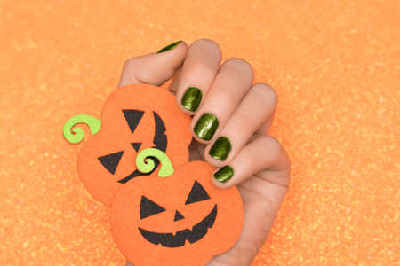 Female hand with green nail design. Glitter green nail polish manicure. Female hold orange paper Halloween pumpkins. Model hand on glitter orange background.の写真素材