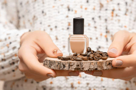 Female hands with beige nail design. Glossy beige nail polish manicure. Model hold beige nail polish bottle with coffee bean on wooden plateの写真素材