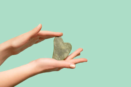 Female model hands with green jade gua sha scraper on green background. Spa body care tool. Beauty care gua sha stone scraper. copy spaceの写真素材