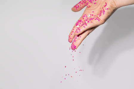 Small pink heart shape sequins fall from young female hand on gray background. Valentine's day concept. Female hand pour pink heart shape sequins on gray background.の写真素材