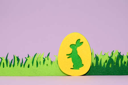 Green easter egg with yellow bunny and fabric green handmade grass on a light purple background. Frame for Easter greeting card with green diy grass and bunnyの写真素材