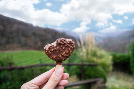 Female hand holding bited chocolate ice cream with nuts on light nature background. Natural ice cream in female hand outdoorの写真素材