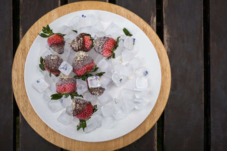 Fresh strawberries dip in chocolate dip with nuts on ice plate. Cold fresh strawberry in milk chocolate. Berries in chocolate snack on wooden backgroundの写真素材