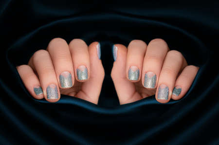 Female hands with silver nail design. Glitter silver nail polish manicure. Female model hands with perfect manicure on dark blue fabric background. Copy space.の写真素材