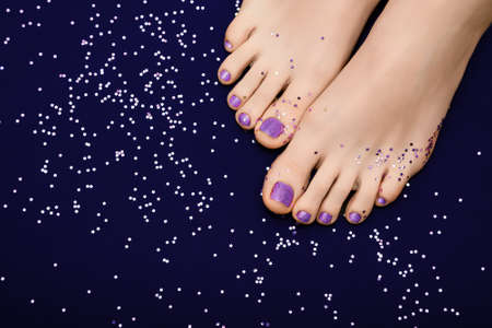 Female feet with purple nail polish pedicure. Woman legs with glitter purple nail design on dark purple fabric background with purple star paillettesの写真素材
