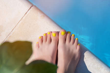 Female with perfect yellow pedicure on a pool side. Vacation pericure. Female feet with yellow pedicure. Female with yellow pedicure stand near blue swimming pool water.の写真素材