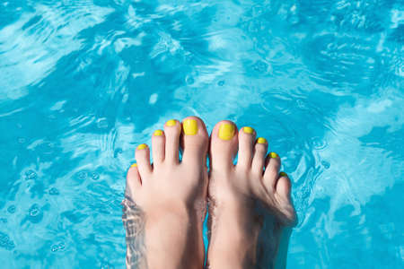 Female with perfect yellow pedicure over a pool with water splash. Vacation pericure. Female bare feet with beautifull yellow pedicure in a blue swimming pool waterの写真素材