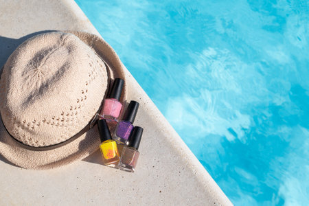 Nail polish bottles: purple, yellow, brown and pink with hat on a pools edge. Nail polish bottles near blue pool water. Vacation manicure nail polish concept. Summer nail polish set with copy space. High quality photo.の写真素材