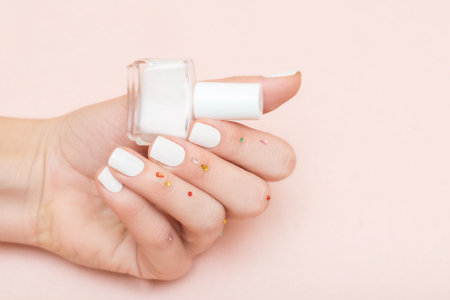 Female hand with white nail design. Glitter white nail polish manicure. Female model hand decorated with red gems with perfect manicure hold white nail polish bottle on pink background. Copy space. Place for text.の写真素材