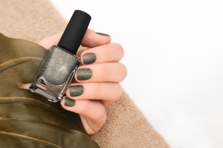 Female hand with brown nail design. Glitter brown nail polish manicure. Female model hand with perfect manicure hold brown nail polish bottle on white background with tropical leaf. Copy space. Place for text.の写真素材