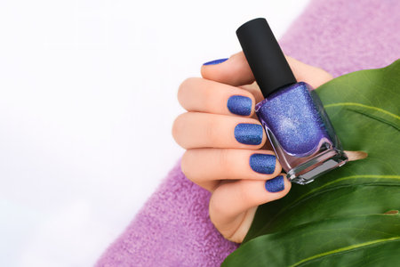 Female hand with purple nail design. Purple glitter nail polish manicure. Female model hand with perfect manicure hold purple nail polish bottle on white background with tropical leaf. Copy space. Place for text.の写真素材