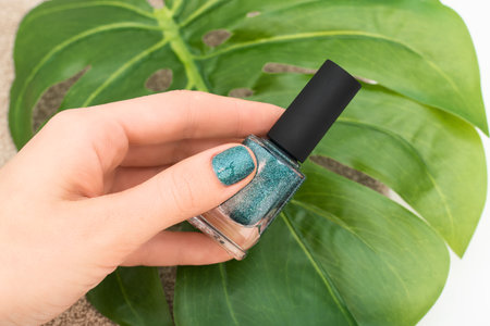 Female hand with blue nail design. Blue glitter nail polish manicure. Female model hand with perfect manicure hold blue nail polish bottle on white background with tropical leaf. Copy space. Place for text.の写真素材