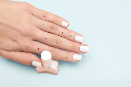 Female hand with white nail design. Glitter white nail polish manicure. Female model hand decorated with red gems with perfect manicure hold white nail polish bottle on blue background. Copy space. Place for text.の写真素材