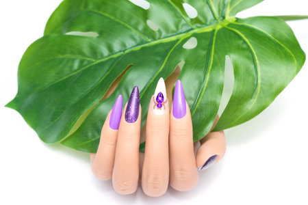 Female hand with vacation stiletto nail design. Glitter purple nail polish manicure with rhinestones spider nail art. Female model hand hold tropic leaf on white background. Copy space. Place for textの写真素材
