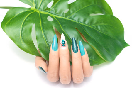 Female hand with vacation stiletto nail design. Glitter cyan nail polish manicure with rhinestones spider nail art. Female model hand hold tropic leaf on white background. Copy space. Place for text.の写真素材