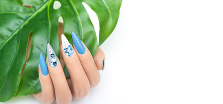 Female hand with vacation stiletto nail design. Glitter blue nail polish manicure with rhinestones and glitter nail art. Female model hand hold tropic leaf on white background. Copy spaceの写真素材