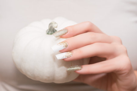 Female hand with long nail design. Glitter gold and white nail polish manicure. Female hand with perfect manicure hold pumpkin.の写真素材