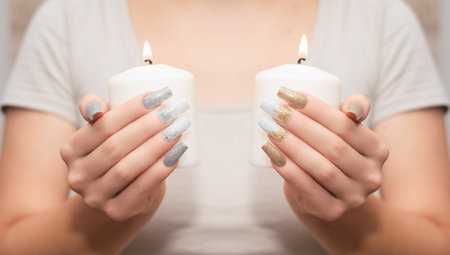 Female hands with long nail design. Glitter gold and white nail polish manicure. Female hands with perfect manicure hold candle.の写真素材