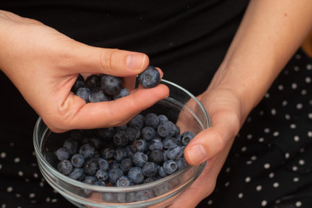Fresh Blueberries in a Glass Bowl in Female Hands. Picking ripe blueberries from a glass bowlの写真素材
