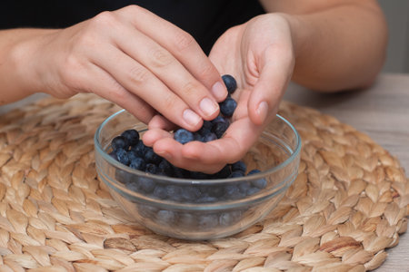 Fresh Blueberries in a Glass Bowl in Female Hands. Picking ripe blueberries from a glass bowlの写真素材