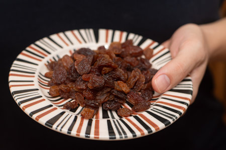 Close-up shot of a person's hands holding a small striped plate filled with juicy, dark raisinsの写真素材
