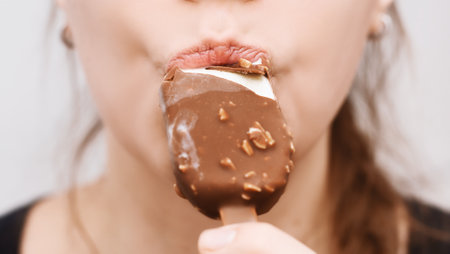 Close-up of Woman Eating Chocolate Ice Cream. Close-up of a woman's lips biting into a chocolate-covered ice cream with nuts on a stickの写真素材