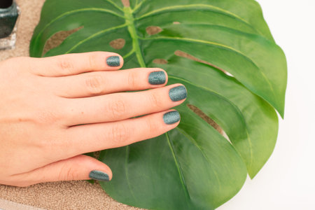 Green Glitter Nail Polish on Female Hand and leaf on background. Close-up of a hand with green glitter nail polish against a green leaf backgroundの写真素材
