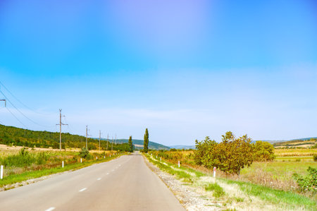 A long, straight rural road with power lines running alongside it, surrounded by fields and trees, under a clear blue skyの写真素材