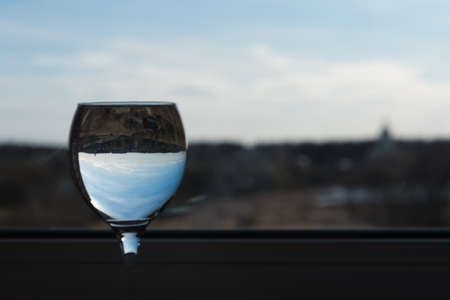 A glass of water on a thin leg stands on the windowsill. Interesting refraction of the view from the window. Sky and clouds in a glass.の写真素材