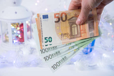 Hand with euro bills. The background consists of light garlands and Christmas toys. Concept of spending for Christmas and New Year. Purchase of New Year's attributes, purchase of Christmas decorations. Christmas lights expenses. Finance.の写真素材