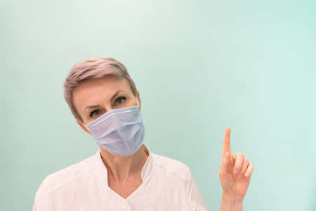 The medic points his finger at a place free for text or images. Doctor, blond woman with short hair. A doctor wearing a disposable medical maskの写真素材