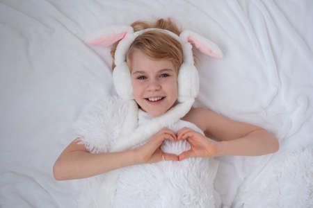 Beautiful little girl plays with rabbit ears. Easter concept. View from above.The child folded the shape of a heart from his fingers. A child on a bed with a fluffy white blanket. Nice and cute picture.の写真素材