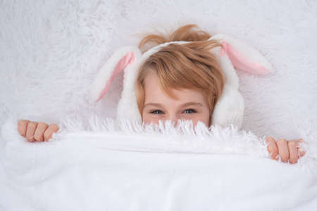 A cute baby appears from under a fluffy white blanket. Teenage girl with rabbit ears. Easter concept. Surpriseの写真素材