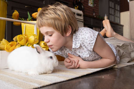 A teenage girl lies on a warm floor in the kitchen. The child kisses the white rabbit and smiles....