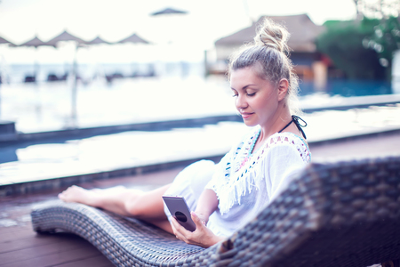 People, technology, travel, tourism and summer concept - happy young woman in bikini swimsuit using smatphone over infinity pool at sea side backgroundの写真素材