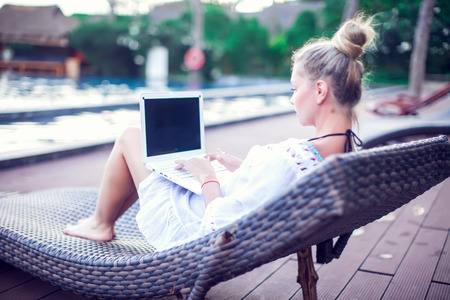 Back view of young woman doing remote work at modern laptop while resting on sun bed near blue swimming pool enjoying summer vacationの写真素材