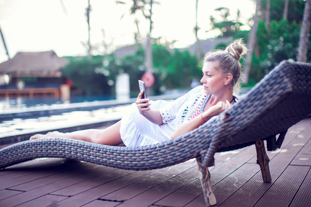 People, technology, travel, tourism and summer concept - happy young woman in bikini swimsuit using smatphone over infinity pool at sea side backgroundの写真素材