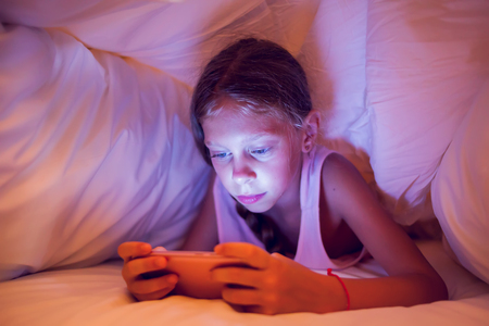 Happy Girl Looking At Mobile Phone While Lying Under Blanket On The Bedの写真素材