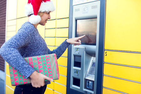 Woman with santa christmas hat client using automated self service post terminal machine or locker to deposit the parcel for storageの写真素材