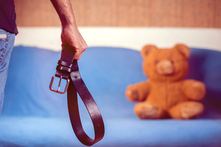 Domestic violence: male hand with belt on the background of toy bear. Abused child, aggression in the family conceptの写真素材