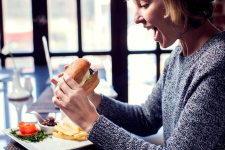 Happy woman is eating burger and smiling while spending time in cafeの写真素材