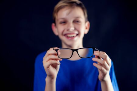 A portrait of a teen boy holds eyeglasses in hands in front of dark background. Children, ophthalmology and healthcare conceptの写真素材