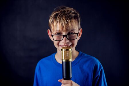 A portrait of a teen boy with eyeglasses holds microphone in front of dark background. Children,...