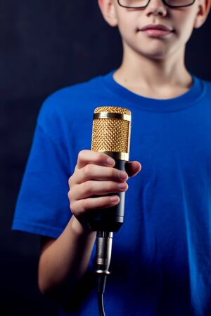 A portrait of a teen boy with eyeglasses holds microphone in front of dark background. Children, talent and entertainment conceptの写真素材