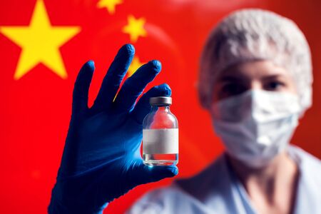 A doctor in blue medical gloves holding a bottle with vaccine in front of flag of China. Close up shot.の写真素材